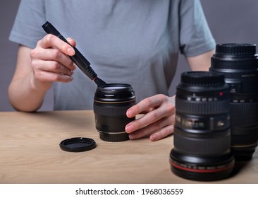 Female photographer cleaning camera lens with professional brush, care about equipment, closeup, removing dust at desk.