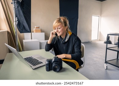 Female photographer smiling while talking on the phone in her studio with a laptop and camera on the desk - Powered by Shutterstock