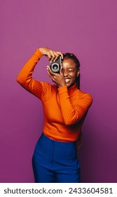 Female photographer stands with confidence and a smile, capturing a fun moment with her digital camera. Vibrant purple background  cheerful smile. Casual style and braided hairstyle add to her lifest