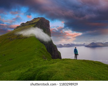 Female photographer stands on the edge of a cliff and watches the spectacular blue and orange evening sky. Stunning view of the magnificent green cliffs and mountains in the picturesque Faroe Islands.