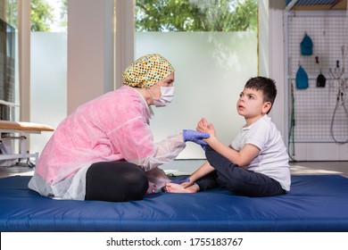 Female Physical Therapist With Disabled Child On Blue Mattress Doing Rehabilitation Exercise. She Wears Coronavirus Protection, Lilac Latex Gloves, White And Pink Suit, Cap And Protective Mask.