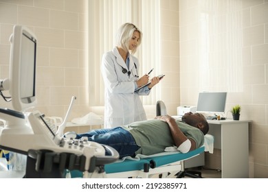 Female Physician In White Coat Taking Notes While Talking To Young Black Male Patient With Bandage On Arm After Venipuncture Lying On Couch In Doctors Office