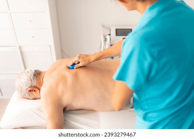 A female physiotherapist massages the back of an older man with a massage tool, a finger protector, during a manual therapy session in a clinic - Powered by Shutterstock