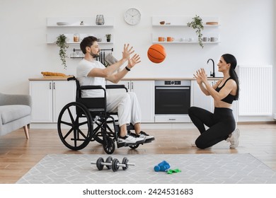 Female physiotherapist or wife throws basketball to young man in wheelchair doing workout exercises at home. Senior male patient undergoing rehabilitation after illness or injury. - Powered by Shutterstock