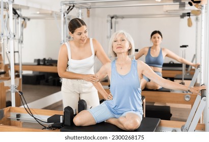 Female pilates instructor helping elderly woman doing exercises on reformer apparatus at gym - Shutterstock ID 2300268175