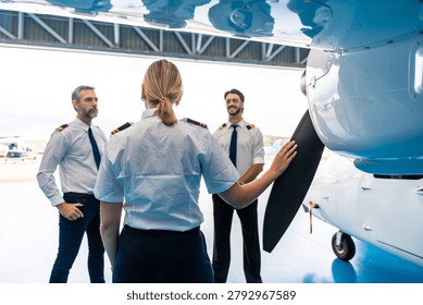 Female pilot touching propeller while discussing flight plans and aviation with two male pilots standing in a professional hangar setting, representing teamwork and training