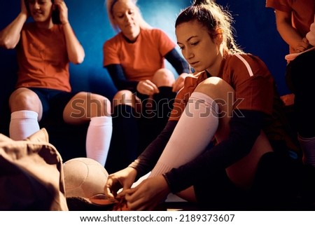 Female player tying soccer cleats while getting ready with her team in dressing room.