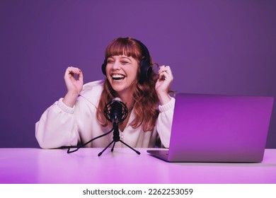 Female podcaster laughing while hosting a live broadcast in a studio. Happy young woman recording an audio show in neon purple light. Woman creating content for her internet podcast. - Shutterstock ID 2262253059