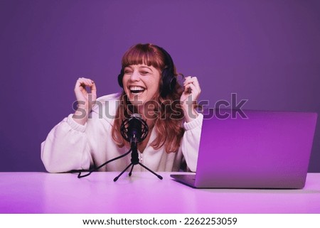 Female podcaster laughing while hosting a live broadcast in a studio. Happy young woman recording an audio show in neon purple light. Woman creating content for her internet podcast.