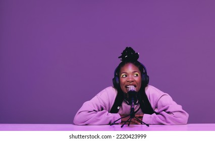 Female podcaster speaking into a microphone while sitting against a purple background. Happy woman recording a live audio broadcast in a studio. Young woman creating content for her internet podcast. - Powered by Shutterstock