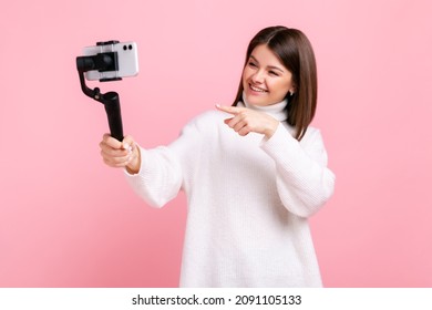 Female Pointing To Cell Phone Camera With Smile, Streaming, Talking With Followers, Uses Steadicam, Wearing White Casual Style Sweater. Indoor Studio Shot Isolated On Pink Background.