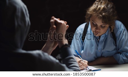 Female police officer interrogating criminal in handcuffs at desk in dark room. Woman cop questioning suspect and writing down information at police department