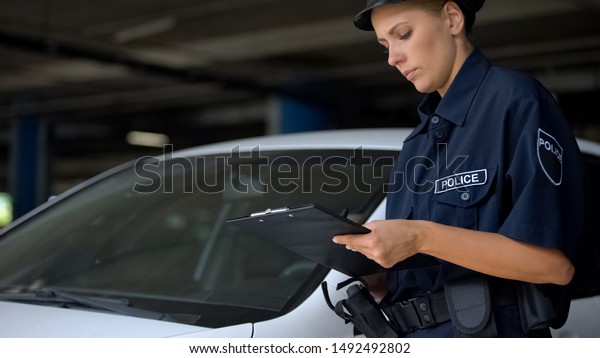 Female police officer writing traffic ticket for\
parking violation near\
car