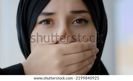 Female portrait indoors Islamic Arabian girl sad afraid Muslim woman Arab lady in black hijab looking at camera covers mouth with hands quiet silence discrimination stop talking oppression of women