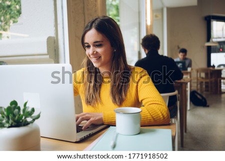 Female positive Caucasian freelancer working on web design project via modern gadget in public area in bar connected to wifi and drinking coffee