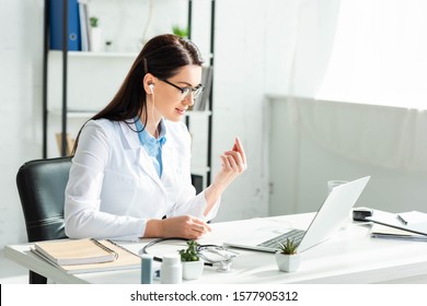 Female Positive Doctor In Earphones Having Online Consultation With Patient On Laptop In Clinic Office 