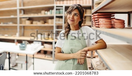 Female potter looking away thoughtfully while standing next to her handmade ceramic products in her shop. Creative young craftswoman running a successful small business.
