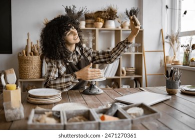 Female potter recording a pottery tutorial on her smartphone while working on a piece in her studio. Cozy, creative workspace with shelves and supplies in the background. - Powered by Shutterstock