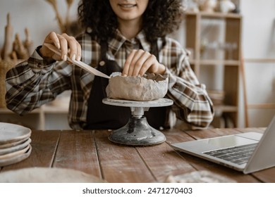 Female potter sculpting clay on spinning wheel in ceramics studio with laptop. Focus on hands and artwork. - Powered by Shutterstock