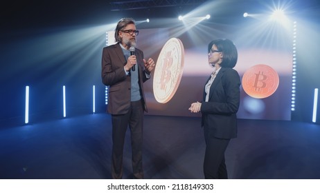 A Female Presenter In A Suit Interviewing A Cryptocurrency Coach On Stage In Front Of An LED Screen With A 3D Object In A Room, With Spotlights During Investment Event