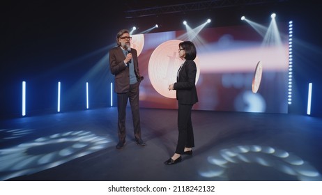 A Female Presenter In A Suit Interviewing A Cryptocurrency Coach On Stage In Front Of An LED Screen With A 3D Object In A Room, With Spotlights During Investment Event