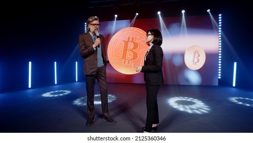 A Female Presenter In A Suit Interviewing A Cryptocurrency Coach On Stage In Front Of An LED Screen With A 3D Object In A Room, With Spotlights During Investment Event