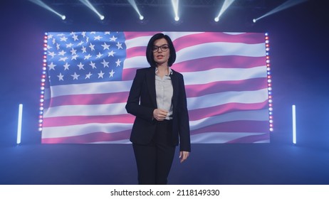 Female Presenter In Suit On Stage Talking About The Latest Political And Public News Of The United States On TV Broadcast In The Modern Studio, With Spotlights Near The American Flag On The LED Screen