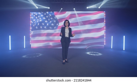 Female Presenter In Suit On Stage Talking About The Latest Political And Public News Of The United States On TV Broadcast In The Modern Studio, With Spotlights Near The American Flag On The LED Screen