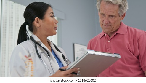 Female Primary Care Physician Taking Medical History From Senior Caucasian Male Patient. Japanese Woman Doctor Talking To Aging Man And Making Notes