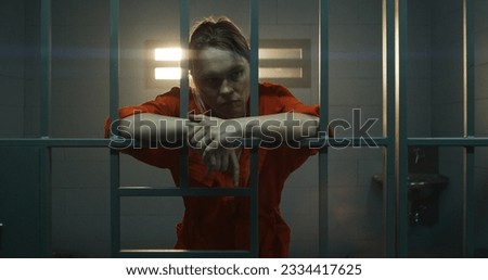 Female prisoner in orange uniform holds hands on metal bars, walks, looks at barred window in jail cell. Woman serves imprisonment term for crime in prison. Depressed inmate in correctional facility.