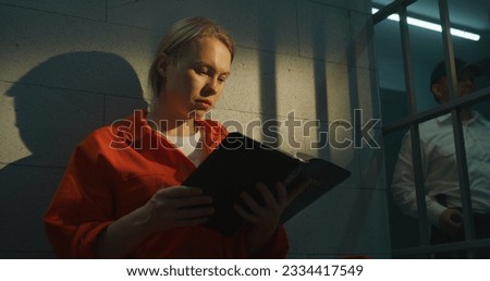 Female prisoner in orange uniform sits on bed in prison cell, reads Bible. Prison officer walks the corridor. Woman criminal serves imprisonment term for crime in jail or correctional facility.