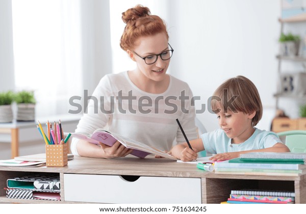 Female private tutor helping young student\
with homework at desk in bright child\'s\
room