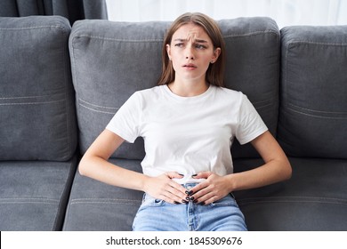 Female problems. Adult woman is sitting on a sofa at home and touching her lower stomach while suffering from cramps. Stock photo - Shutterstock ID 1845309676