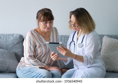 Female Professional Doctor Showing Medical Test Result Explaining Prescription Using Digital Tablet App Visiting Senior Woman Patient At Home Sitting On Sofa. Elderly People Healthcare Tech Concept.