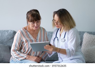 Female Professional Doctor Showing Medical Test Result Explaining Prescription Using Digital Tablet App Visiting Senior Woman Patient At Home Sitting On Sofa. Elderly People Healthcare Tech Concept.