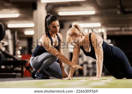 A female professional trainer is training a strong woman who is doing pushups in a gym.