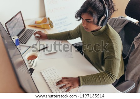 Female programmer coding on a desktop computer and laptop at her desk in office. Young woman in casuals wearing headphones working on a laptop and desktop computer.