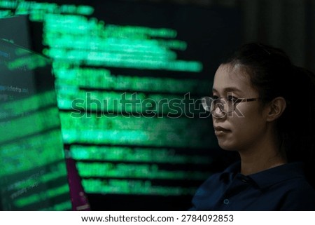 A female programmer working in a dark office environment while monitoring cybersecurity data and digital code on a computer screen. The scene represents software development, cyber technology, data analysis, and information security in a modern technology workspace.