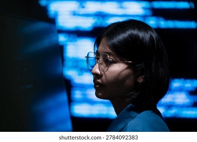 A female programmer working on a computer with illuminated digital information screens in a dark technology office. The image represents software engineering, cybersecurity monitoring, and modern digital workspace environments.
