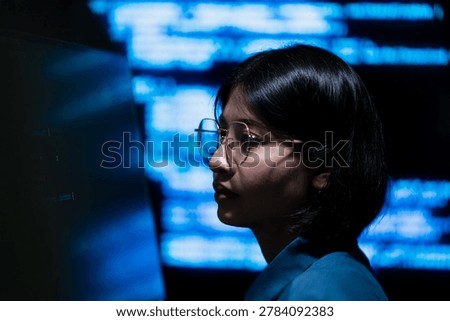 A female programmer working on a computer with illuminated digital information screens in a dark technology office. The image represents software engineering, cybersecurity monitoring, and modern digital workspace environments.