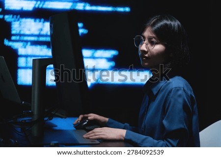 A female programmer working on a computer with illuminated digital information screens in a dark technology office. The image represents software engineering, cybersecurity monitoring, and modern digital workspace environments.