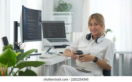 Female programmers sitting front of laptop and large curved computer monitor with programming code on screen and smiling to camera.
