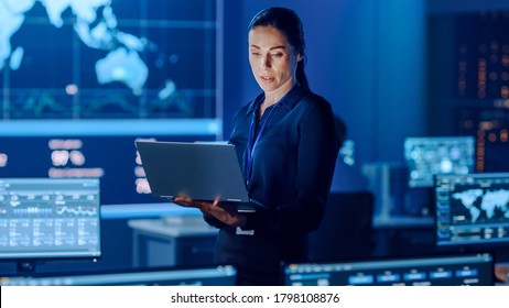 Female Project Leader is Standing with Laptop Computer and Checking Work Data. Science Engineers Work Around Her. Telecommunications Control Monitoring Room with Neural Network on Servers.