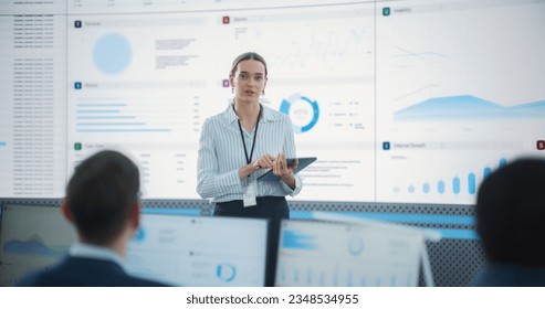 Female Project Manager With Tablet Computer Is Giving Presentation To Diverse Employees In Front of Big Digital Screen In Monitoring Room. Team Of Data Scientists Listening To New Objectives, Tasks.