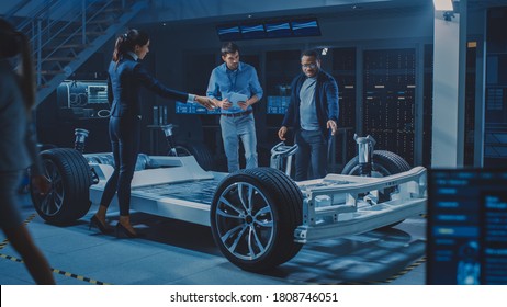 Female Project Manager Talks about Electric Car Prototype with Her Team of Engineers, Showing Chassis, Explaining Sustainability and Ecological Efficiency of the Engine - Powered by Shutterstock