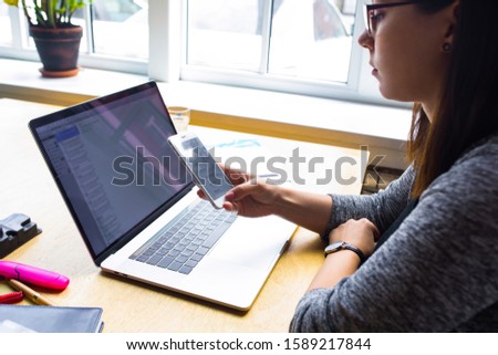 Female proud digital projects development using messenger on mobile phone while sitting with laptop computer in office interior. Hipster girl checking e-mail on cell telephone during work on netbook 