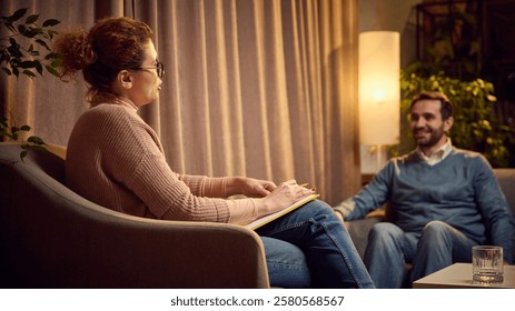 Female psychologist with notes explaining something to a male client seated on a couch in a warm, cozy therapy space. Concept of psychology, assistance, therapy