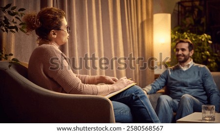 Female psychologist with notes explaining something to a male client seated on a couch in a warm, cozy therapy space. Concept of psychology, assistance, therapy