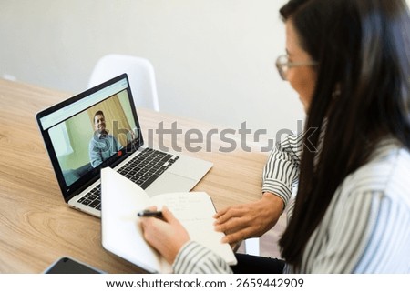 Female psychologist taking notes while attending an online therapy session with her patient via video call on a laptop computer