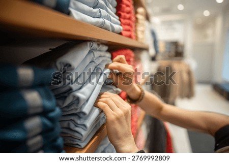 Female purchasing new basic clothing at garment fashion showroom, woman looking through stack of t-shirts on shelf display, examinees quality of material, fabric and preferable size of clothing item.